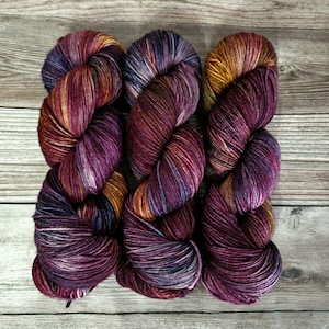 May include: Three skeins of hand-dyed yarn in shades of purple, burgundy, gold, and blue. The yarn is arranged on a wooden surface, showcasing the colour variations and textures. Ideal for knitting or crochet projects.