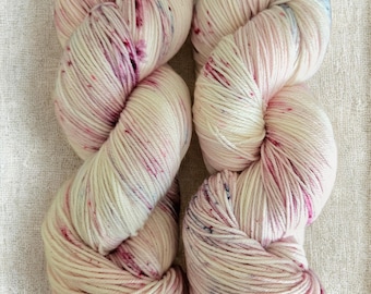 May include: Two skeins of yarn in a cream color with pink and purple speckles. The yarn is tightly wound, showing the texture of the fibers. The yarn is suitable for knitting or crocheting projects.