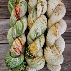May include: Three skeins of hand-dyed yarn in a natural color palette. The yarn is a creamy white base with splashes of green, orange, and brown. The yarn is arranged in a twisted, coiled shape, perfect for knitting or crocheting projects.