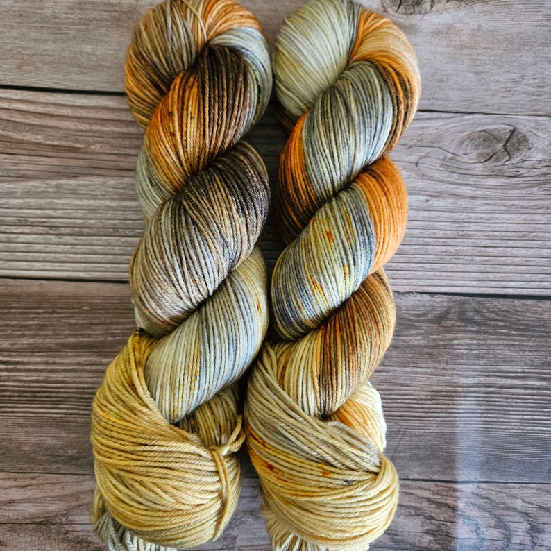 May include: Two skeins of hand-dyed yarn in a variegated colourway. The yarn features shades of brown, orange, yellow, and grey. The yarn is twisted into a rope-like shape, with the colours blending together. The background is a wooden surface.