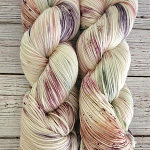 May include: Two skeins of hand-dyed yarn in a variegated colourway with shades of cream, pink, purple, and green. The yarn is a soft, fluffy texture and is perfect for knitting or crocheting.