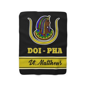 May include: Black fleece blanket with a gold and black design. The design features an Egyptian queen with a gold crown and a gold ankh symbol. The text "DOI - PHA Dt. Matthews" is printed in gold letters.