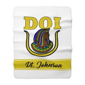 May include: White fleece blanket with a gold and black design. The design features the letters DOI in gold, a stylized image of an Egyptian queen with a gold crown and a gold U-shaped symbol. The text "Dt. Johnson" is written in black cursive on a gold stripe.