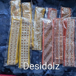 May include: A collection of decorative, embellished ribbons in clear plastic packaging. The ribbons feature gold, silver, and orange-toned rhinestones and beads. The text "Desidolz" is visible on the bottom of the image.