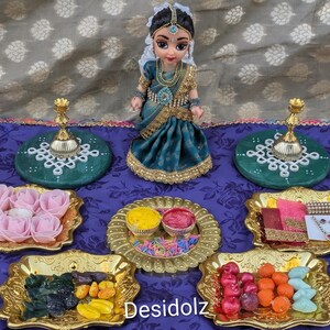 May include: A doll dressed in a teal and gold sari stands on a purple cloth. Surrounding the doll are gold-toned trays holding pink foam roses, yellow and red powders, colorful sprinkles, small sweets, and jewelry. Two green circular stands with gold candle holders are placed behind the doll. The text Desidolz is visible at the bottom.