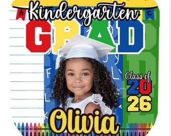 May include: A personalized kindergarten graduation photo featuring a young girl wearing a graduation cap. The design includes the text "Kindergarten GRAD" in bold letters, the name "Olivia", and "Class of 2026". The background has a school theme with crayons and a ruler.