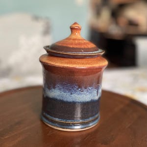 May include: A handcrafted ceramic jar with a lid. The jar features a gradient of colors, transitioning from brown to blue and black. The lid has a small knob on top. The jar is sitting on a wooden surface.