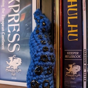May include: A blue crocheted octopus with black buttons for eyes. The octopus is standing in front of three books with the text "Chaossium Inc." and "Keeper Rulebook" on the spines.