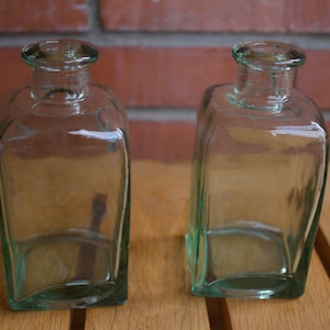 Può includere: Due bottiglie di vetro trasparente con forma quadrata e una leggera sfumatura verde. Le bottiglie sono vuote e sono appoggiate su una superficie di legno.