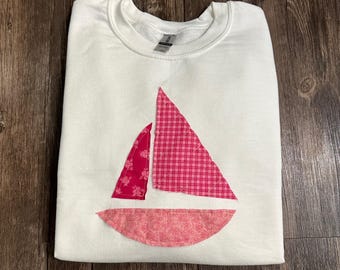 May include: White sweatshirt with a pink and red sailboat applique. The sailboat is made of fabric with a floral pattern and a plaid pattern.