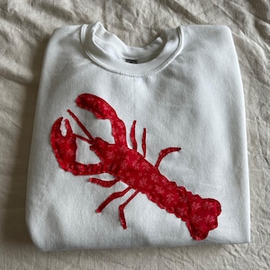 Può includere: Felpa bianca con un'applicazione di aragosta rossa. L'aragosta è realizzata in tessuto con un motivo floreale rosso e bianco.