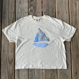 Puede incluir: Una camiseta blanca recortada con una aplicación de barco de vela azul y blanco. El barco de vela está hecho de tela y tiene un patrón floral.