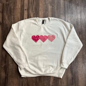 Op de afbeelding: Een wit sweatshirt met drie roze en rode patchwork harten op de voorkant.