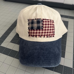 May include: A baseball cap with an off-white crown and a distressed dark blue brim. The front features a rectangular patch made of navy blue plaid and red and white gingham fabric, secured to the cap with visible black cross-stitch embroidery.