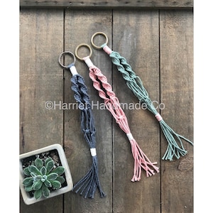 Könnte beinhalten: Drei Makramee-Pflanzenhänger in verschiedenen Farben: Grau, Rosa und Grün. Jeder Hänger hat oben einen Metallring und ist aus einem geknüpften Seildesign gefertigt.