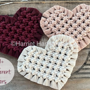 May include: Three handmade macrame heart wall hangings in burgundy, blush pink, and cream colors. The hearts are made with cotton cord and have fringe details. The text "50+ Different Colors" is visible in the image.