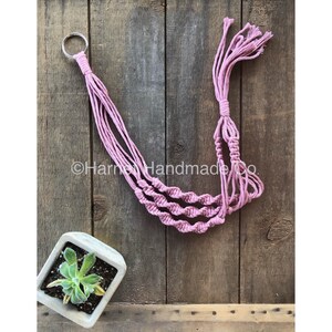 May include: A dusty rose macrame plant hanger with a silver metal ring at the top. The plant hanger has a spiral design and is made of knotted cotton rope. A small square concrete planter with a succulent plant is in the lower left corner.