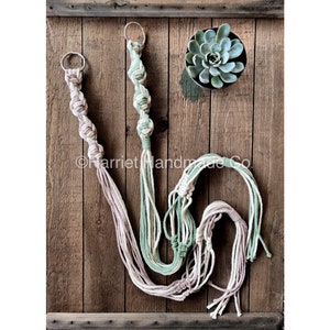 May include: Two macrame plant hangers with a green and white color scheme and a pink and white color scheme. The hangers are made with cotton cord and have a metal ring at the top.