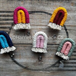 May include: Five colorful rainbow-shaped crocheted badge holders with white fringe. Each holder is attached to a clear plastic badge clip.