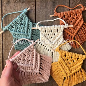 May include: Five small macrame wall hangings with wooden dowels. The hangings are in various colors, including teal, cream, peach, and mustard yellow.