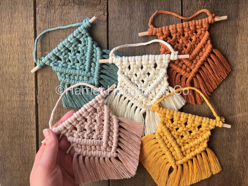 May include: Five small macrame wall hangings with wooden dowels. The hangings are in various colors, including teal, cream, peach, and mustard yellow.