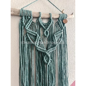 May include: A teal macrame wall hanging with a wood branch at the top. The macrame is in a geometric pattern with a fringe at the bottom. The wood branch has a small wooden tag with the text "Harriet Handmade Co."