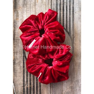 Op de afbeelding: Twee rode fluwelen scrunchies op elkaar gestapeld. De scrunchies zijn gemaakt van zacht, pluche fluweel en hebben een geplooide, gerimpelde look. De scrunchies liggen op een houten achtergrond.