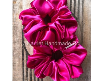 May include: Two bright pink satin scrunchies on a wooden background. The scrunchies are made of soft, silky fabric and are perfect for holding hair in place.