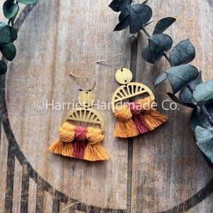 May include: Gold-coloured earrings with a half-circle design and orange and red tassels. The earrings are displayed on a wooden surface with green foliage. The text "Harriet Handmade Co." is visible.