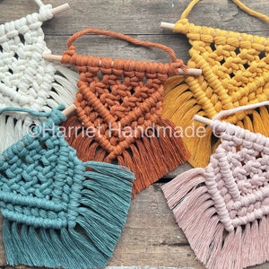 May include: Five small macrame wall hangings with wooden dowels. The hangings are in various colors, including white, orange, yellow, teal, and pink.