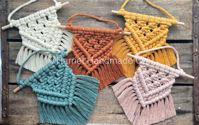 May include: Five small macrame wall hangings with wooden dowels. The hangings are in various colors, including white, orange, yellow, teal, and pink.