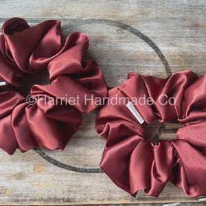 May include: Two burgundy satin scrunchies. The scrunchies are made of a shiny, ruffled fabric. Each scrunchie has a small white tag with the text "Harriet Handmade Co." The scrunchies are on a wooden surface.