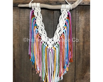 May include: A white macrame wall hanging with a driftwood hanger and rainbow colored yarn tassels.  The macrame is in a V shape with a knot pattern.  The tassels are in shades of pink, orange, yellow, green, blue, and purple.