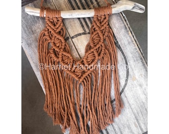 May include: A brown macrame wall hanging with a driftwood branch at the top. The macrame is in a V-shape with fringe hanging down. The background is a wooden surface.