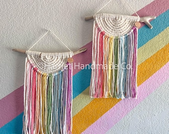May include: Two rainbow macrame wall hangings with a white background and a wooden branch at the top. The macrame is made with a variety of colors, including pink, yellow, green, and blue. The text "Pretty Handmade Co." is visible on the bottom right of the image.