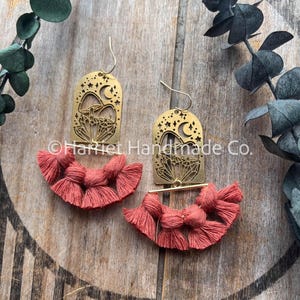May include: Brass earrings with a celestial design, including a crescent moon and stars, combined with coral macrame fringe. The earrings are presented on a wooden surface, with green foliage in the background.
