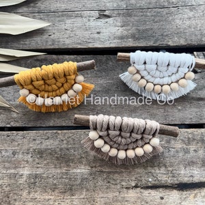 Peut inclure: Trois tentures murales en macramé avec des chevilles en bois et des perles en bois. Les tentures murales sont dans des tons de jaune, blanc et marron.