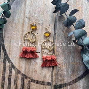 May include: Gold-coloured earrings with amber-coloured square gems and rust-coloured fringe. The earrings feature a sunburst design and are suspended from gold-coloured ear wires. The earrings are displayed on a wooden surface.