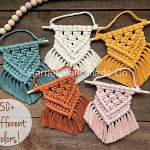 Peut inclure: Cinq petites tentures murales en macramé avec des chevilles en bois et des glands. Les tentures sont de différentes couleurs, notamment turquoise, blanc, jaune, orange et rose. Le texte "50+ Different Colors!" est visible sur l'image.