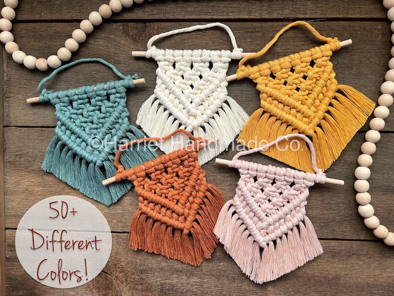 May include: Five small macrame wall hangings with wooden dowels and tassels. The hangings are in various colors, including teal, white, yellow, orange, and pink. The text "50+ Different Colors!" is visible in the image.