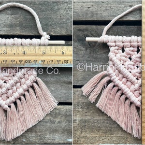 May include: A small pink macrame wall hanging with a wooden dowel and fringe. The hanging is approximately 6 inches tall and 5 inches wide.