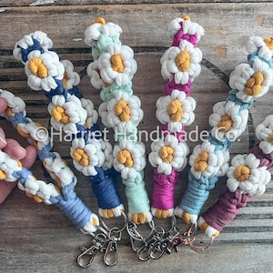 Op de afbeelding: Zes kleurrijke gebreide daisy sleutelhangers met gele centra en witte bloemblaadjes. De sleutelhangers zijn bevestigd aan zilveren sleutelringen.