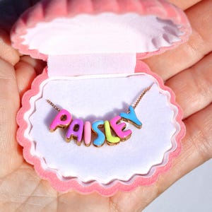 May include: A gold chain necklace featuring colorful enamel letter charms that spell out the name PAISLEY. The letters are in shades of pink, blue, and yellow. The necklace is presented inside a small, pink, shell-shaped jewelry box.
