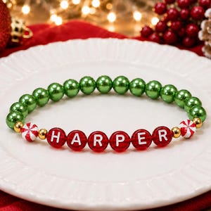 Puede incluir: Una pulsera de cuentas con una hilera de cuentas redondas de color verde met&aacute;lico, seguidas de cuentas rojas que deletrean el nombre HARPER en letras blancas, acentuadas con peque&ntilde;as cuentas espaciadoras doradas y dos cuentas blancas con rayas rojas.