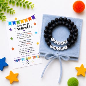 Puede incluir: Dos pulseras de cuentas de lava negra con cuentas de letras blancas que forman LOGAN y DADDY, sobre una bolsa de fieltro azul claro. Una tarjeta blanca presenta banderines coloridos, estrellas y texto sobre el primer día de escuela.