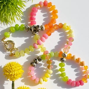 Peut inclure: Un collier de perles colorées avec un fermoir doré. Le collier présente un dégradé de couleurs, notamment des perles roses, oranges, jaunes et vertes. Certaines perles sont mouchetées de noir et de blanc. Le collier est présenté sur une surface blanche.