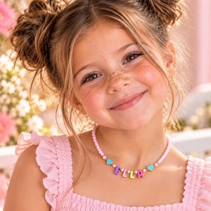 May include: A beaded necklace featuring light pink round beads, small gold-tone spacers, and two turquoise heart charms flanking the name ELLA spelled out in colorful, block-style letter beads in purple, yellow, pink, and orange.