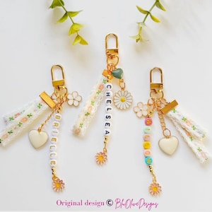 May include: Three personalized keychains with gold hardware, white floral ribbons, and daisy charms. Each keychain features a name spelled out with letter beads, a heart charm, and a flower charm. The keychains are displayed on a white surface with floral accents.