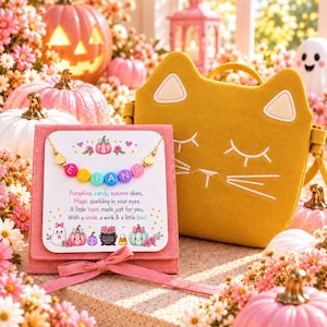 Puede incluir: Un collar dorado con cuentas de letras coloridas que forman el nombre ELIANA sobre una tarjeta blanca con ilustraciones de calabazas y fantasmas, junto a un bolso amarillo mostaza con forma de gato y rasgos bordados.