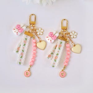 May include: Two personalised keychains with gold-tone clasps. Each keychain has a white bunny charm with a pink bow, a white flower, and a yellow heart. One keychain spells "MADELYN" and the other "SOPHIA" in pink beads. A pink heart charm completes the design.